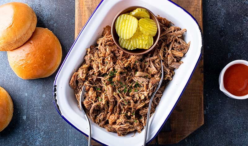 Barbecued pork butt in a white enamel dish with pickles and buns on the side.